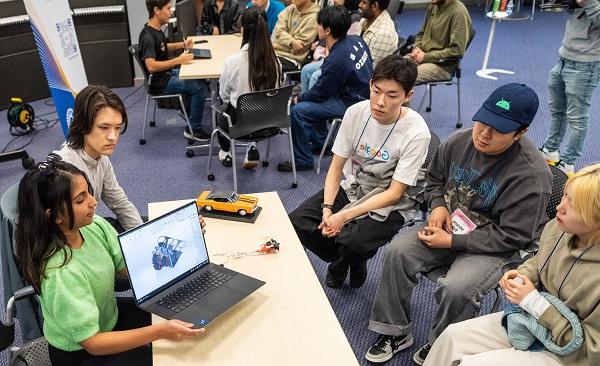 A young woman with dark hair in a greet shirt shows a prototype diagram on her laptop to three young Asian men on the opposite side of a table.  Her male partner sits next to her, with an orange prototype car on the table in front of him.